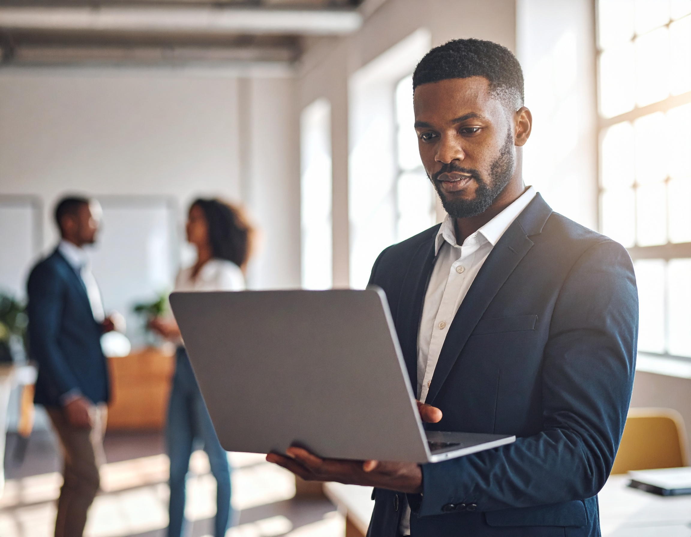 Homem em terno escuro segura um laptop em um escritório iluminado por luz natural, com duas pessoas ao fundo desfocadas em uma discussão. A cena capta um ambiente de trabalho moderno e colaborativo, com foco claro no homem em primeiro plano e uma composição que enfatiza a profundidade do espaço. As janelas grandes permitem a entrada de luz suave, criando uma atmosfera profissional e acolhedora.